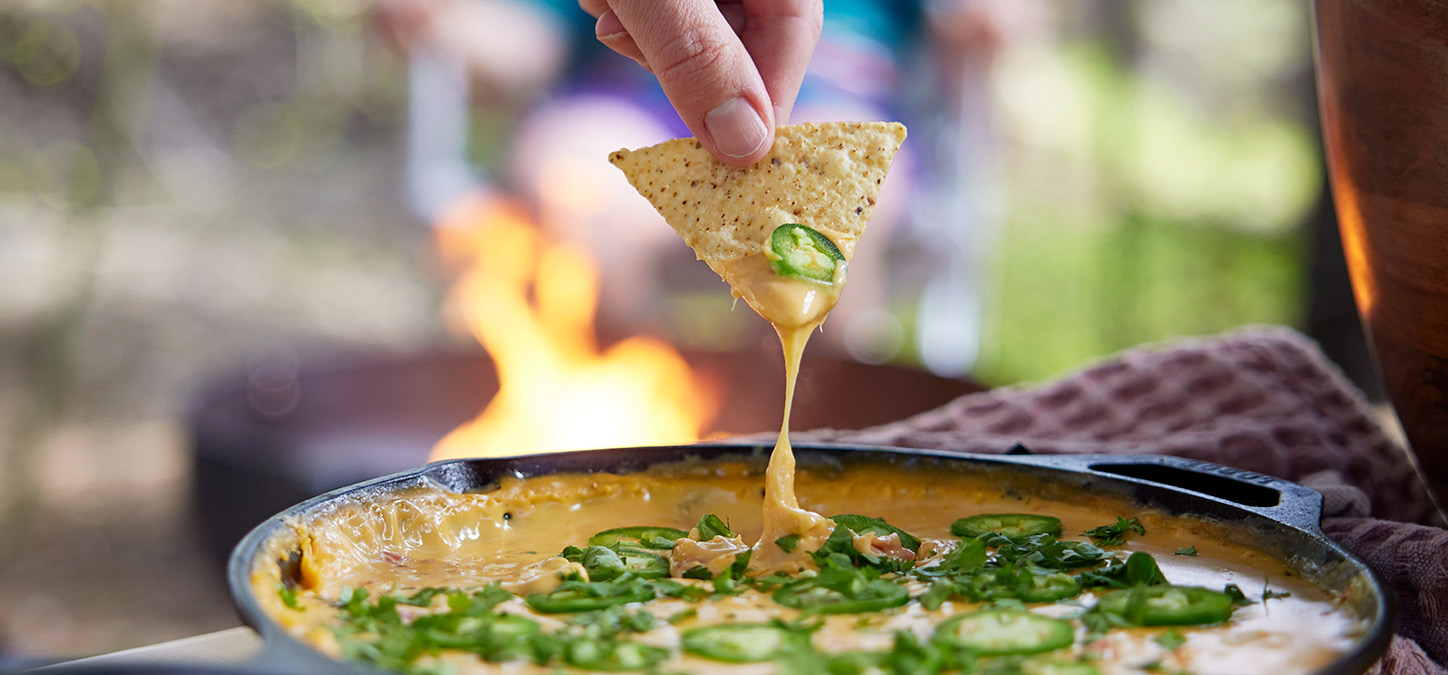 Hand dipping corn chip in spicy queso dip with a Duraflame fire burning in the background