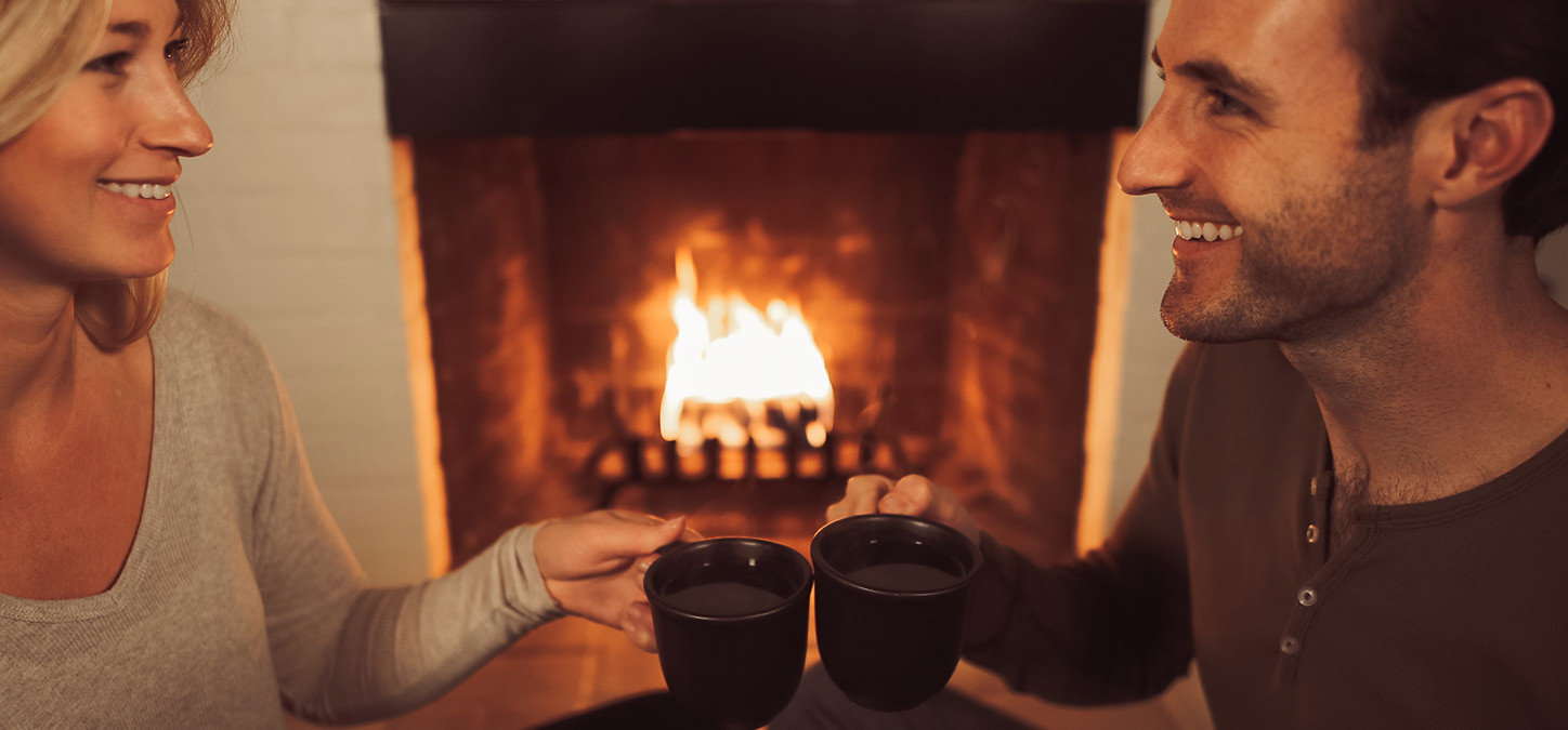 Man and woman smiling, holding mugs of tea with duraflame fire burning in hearth in background