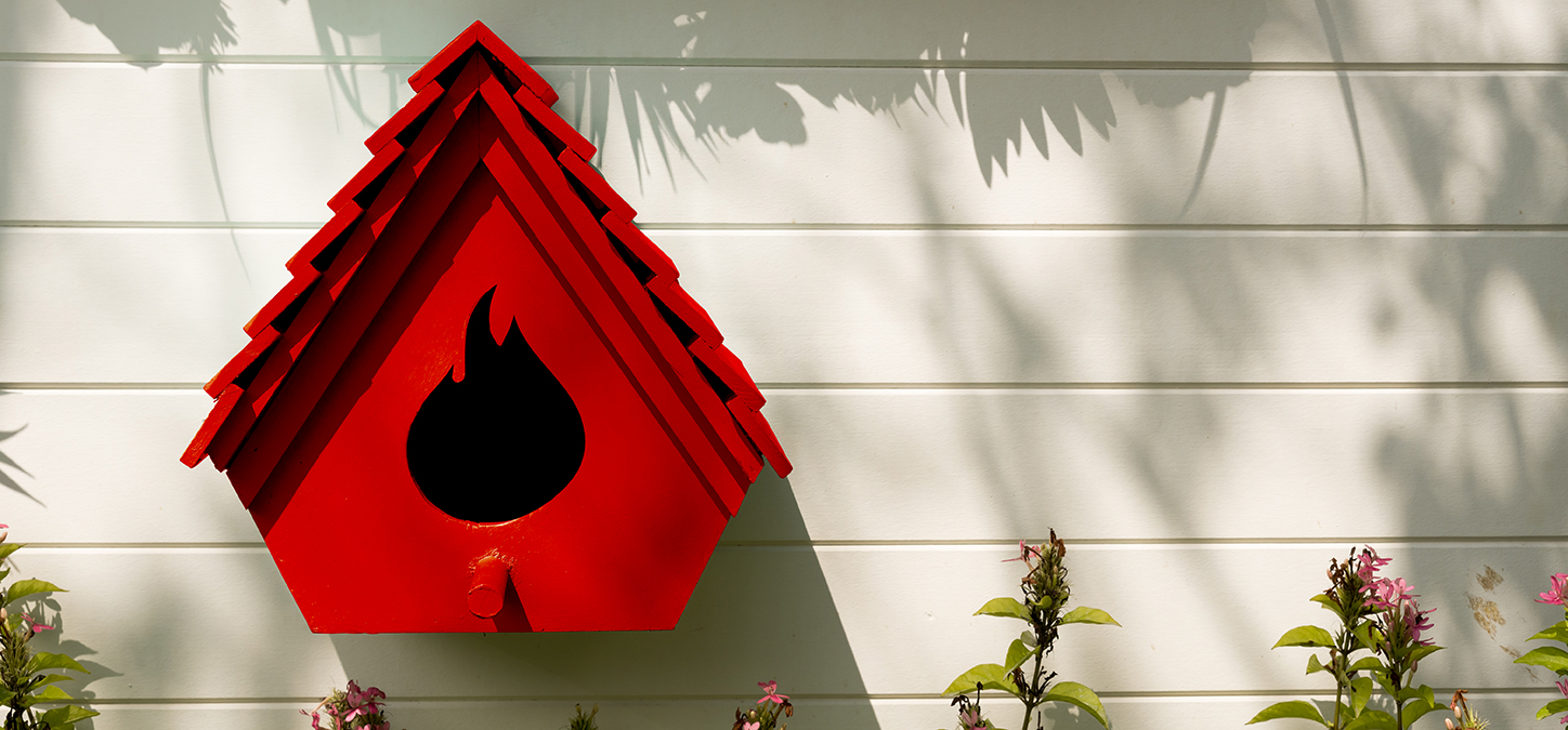 Bright red bird feeder with duraflame brand flame-shaped opening hung outside house above flowers
