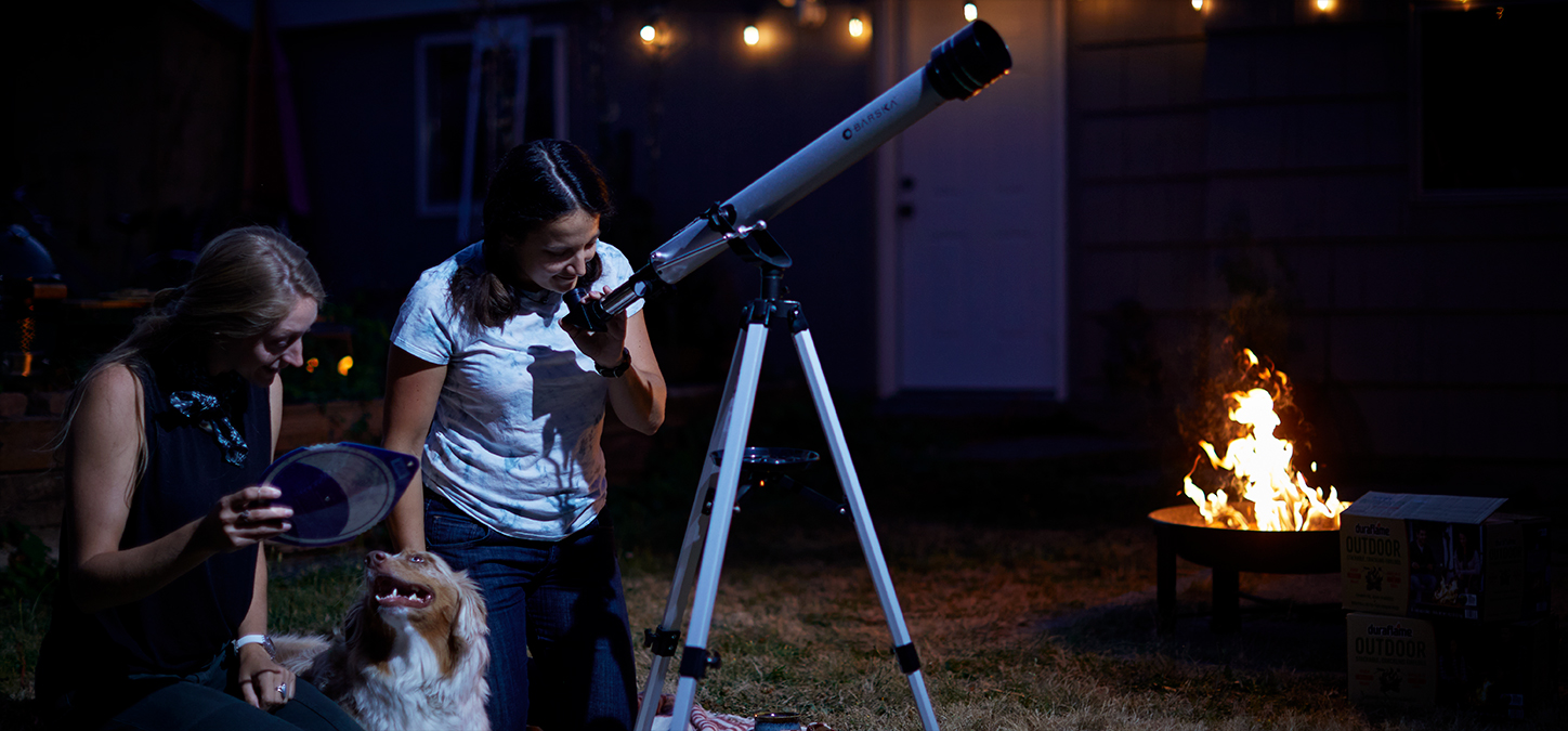 People on blanket with dog stargazing through telescope while fire pit fire burns in background