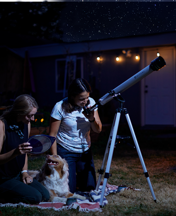 People on blanket with dog stargazing through telescope while fire pit fire burns in background