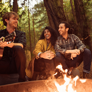 Friends laughing as one plays guitar sitting at a campfire in the woods