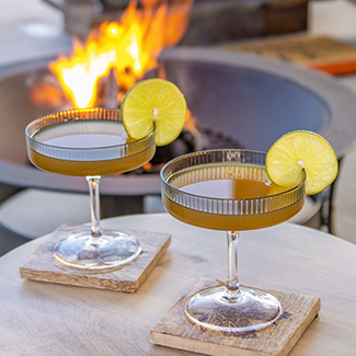 Two cocktails with lime garnish with a duraflame fire burning in an outdoor firepit in the background