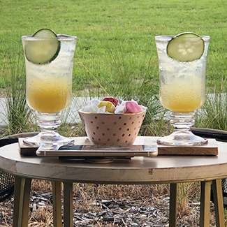 Cold drinks on ice with cucumber garnish on a table with bowl of taffy in the backyard