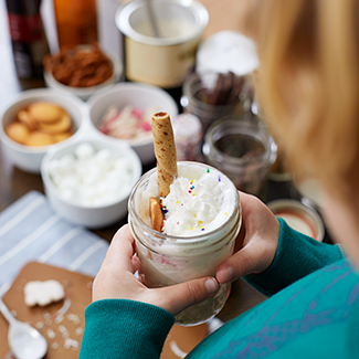 Birds eye view of bowls of sweet toppings and child holding a hot cocoa with whipped cream and cookie on top
