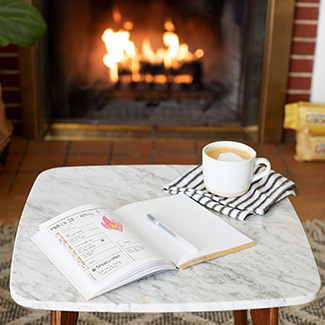 Journal, pen and latte on a table with a duraflame Crackleflame firelog burning in the background
