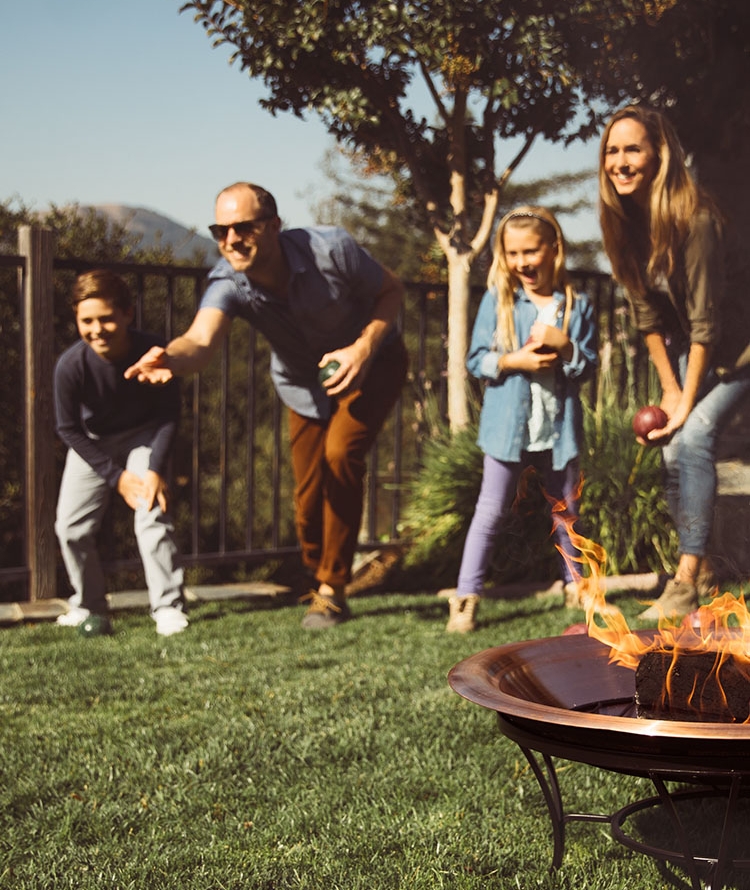 Family playing bocce on backyard lawn with duraflame OUTDOOR firelogs burning in fire pit in foreground