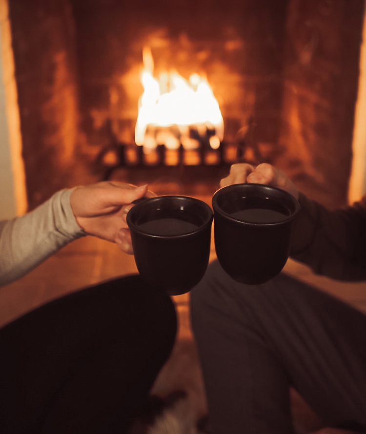 Hands holding mugs of tea with duraflame fire burning in hearth in background