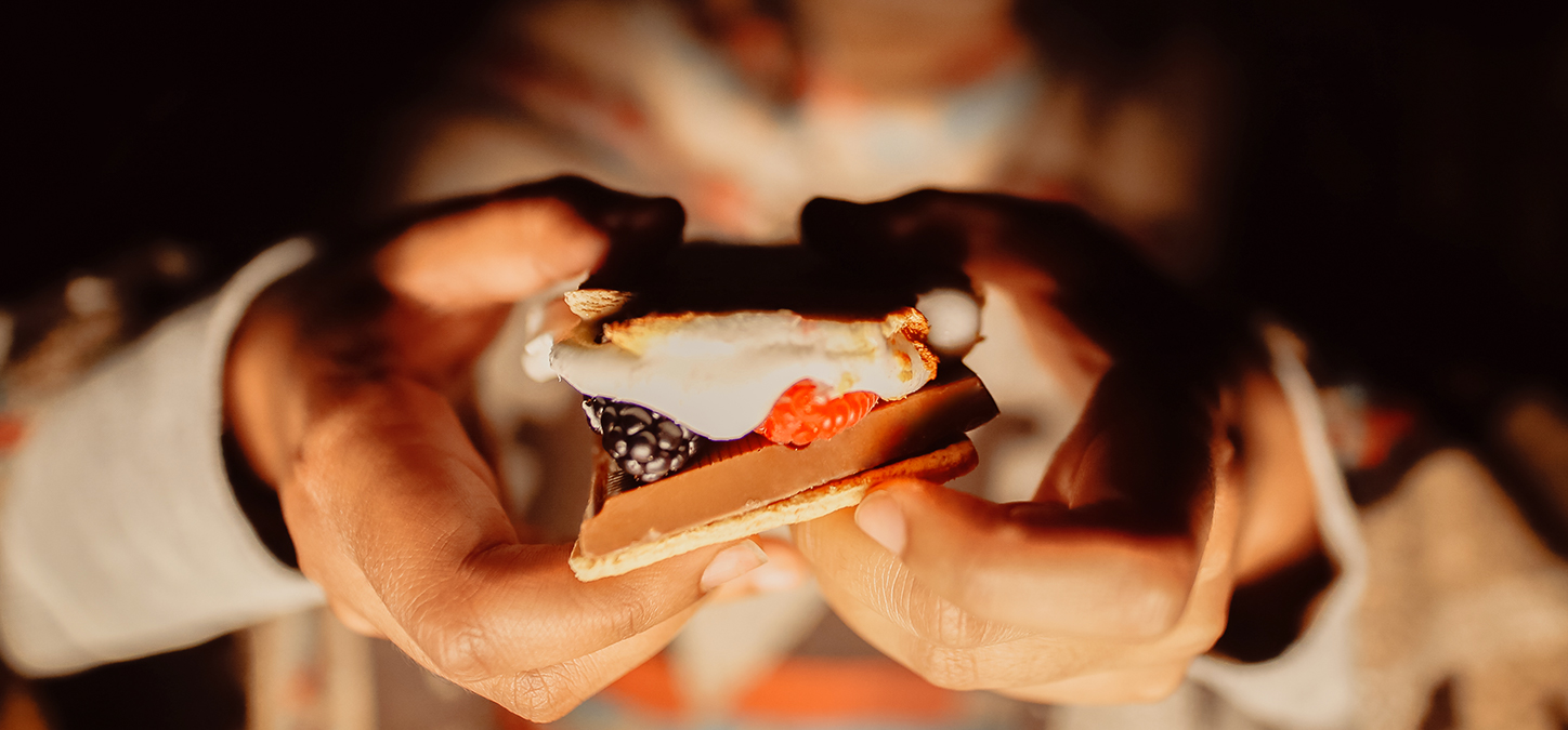 Close up of hands holding s'mores with roasted marshmallow, chocolate and berries.