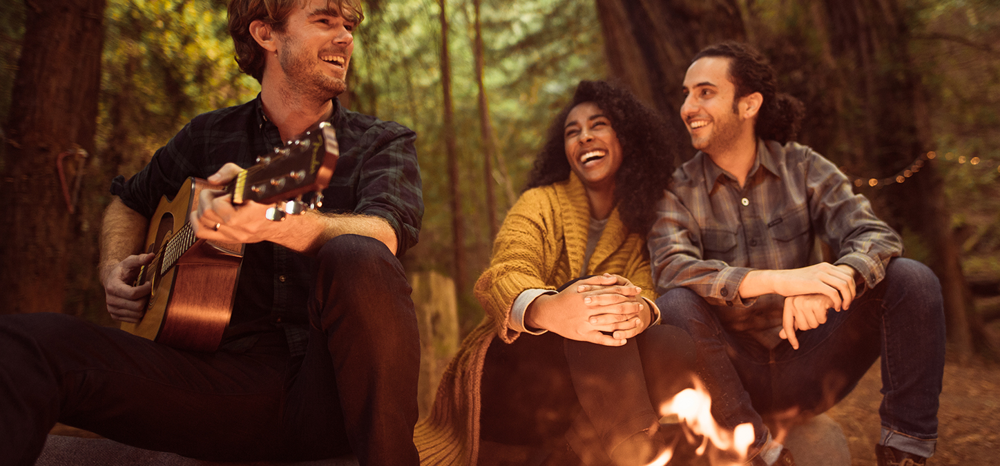 Friends laughing as one plays guitar sitting at a campfire in the woods