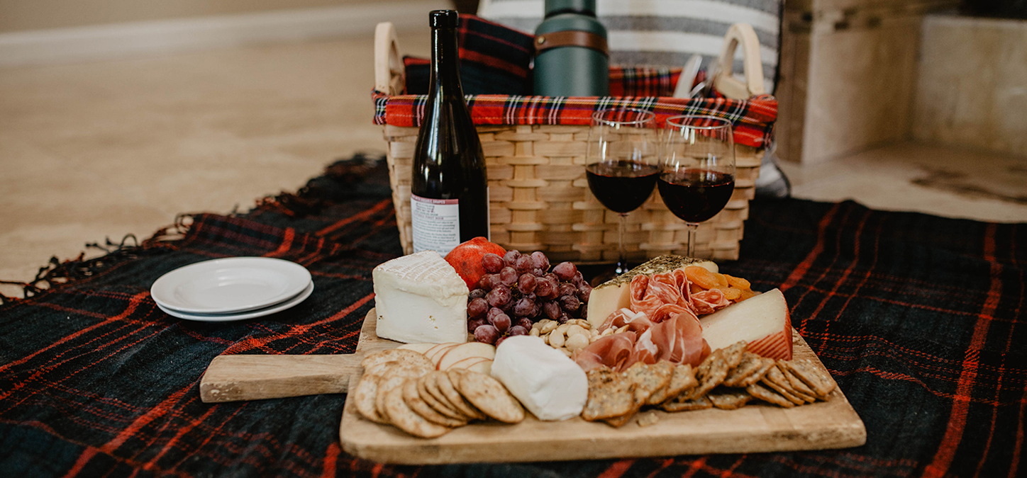 Charcuterie cheeseboard on blanket with wine and picnic Basket in background