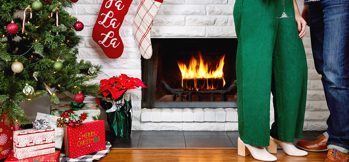 Two people standing next to duraflame fire with stockings hanging and Christmas tree with presents