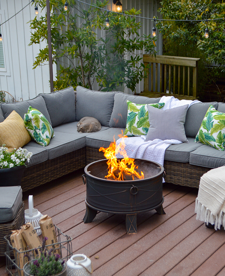 An outdoor seating area with a sofa and cushions, a lit duraflame fire pit, decorative lights above, and a cat resting on the couch.
