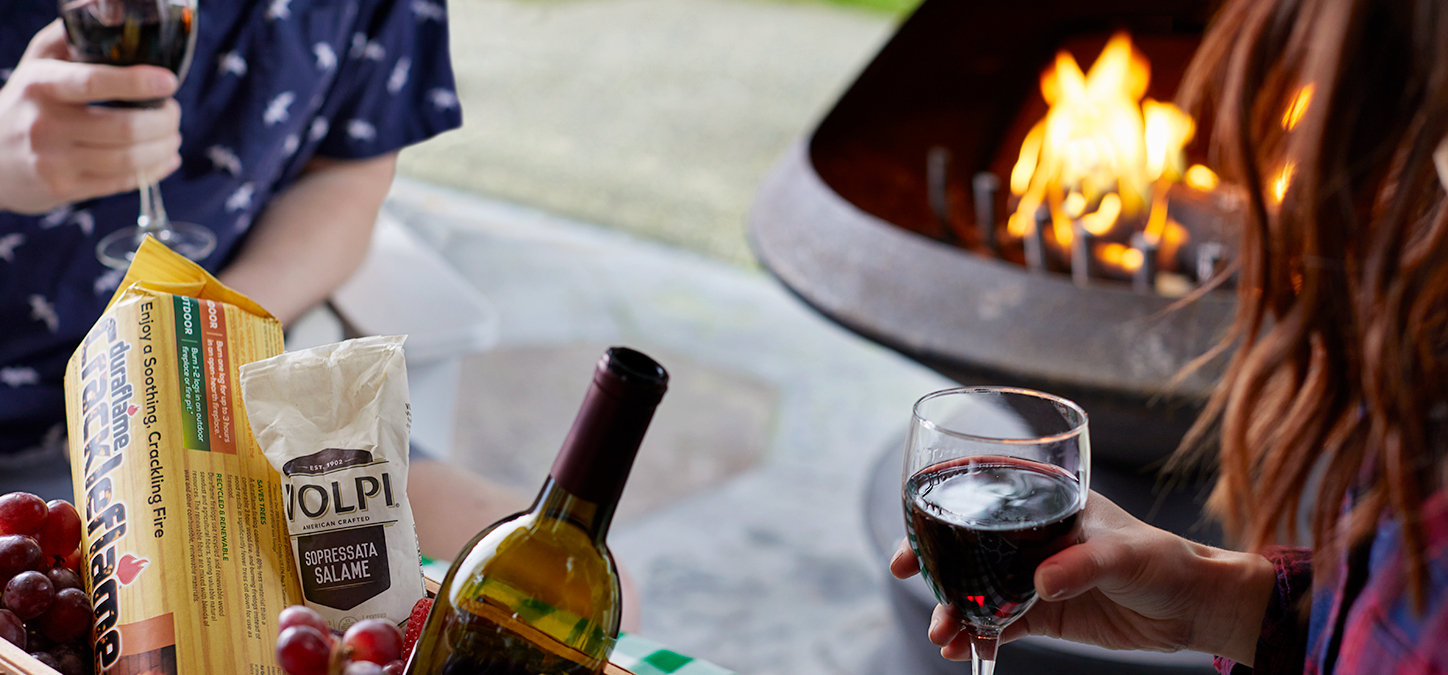 Couple holding wine glasses with picnic basket, enjoying a duraflame Crackleflame fire in a backyard fire pit