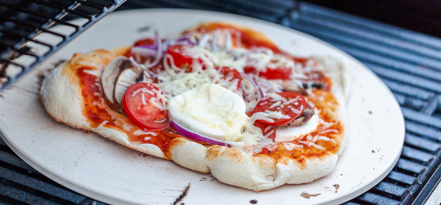 Pizza topped with mushrooms, tomatoes, onions & mozarella on a pizza stone on a grill 