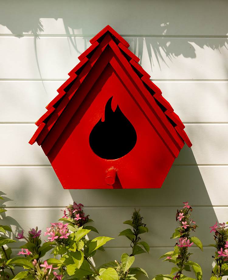 Bright red bird feeder with duraflame brand flame-shaped opening hung outside house above flowers