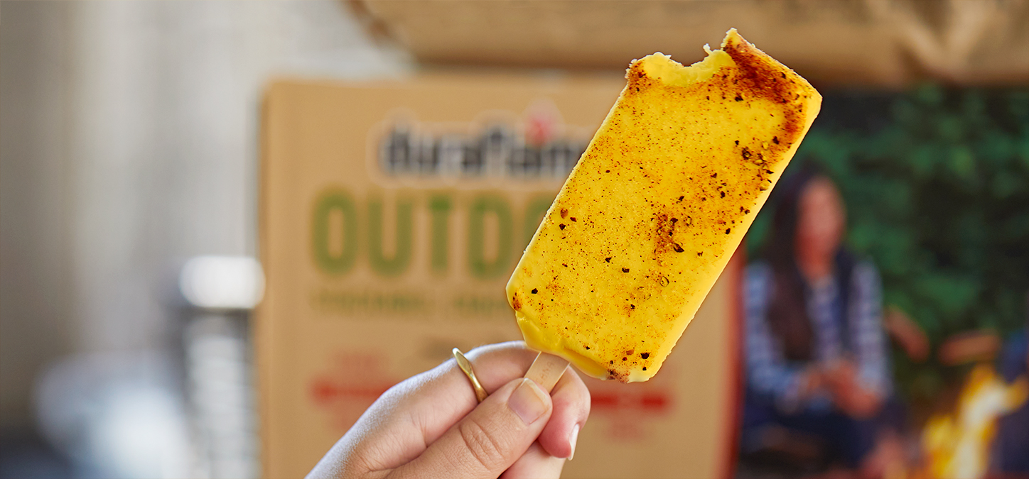 A hand holding a yellow popsicle with red spices over a plate with a  case of duraflame OUTDOOR firelogs in the background
