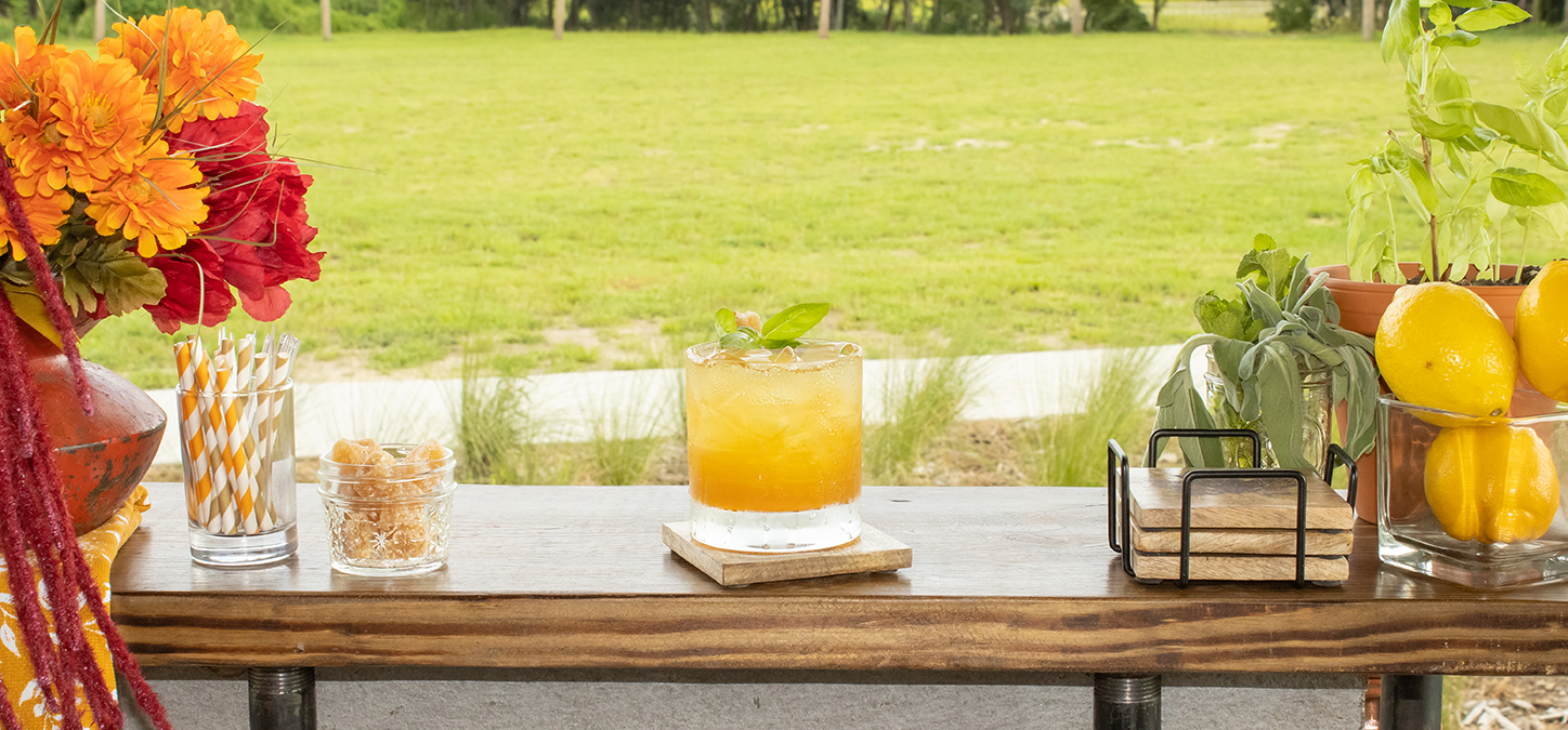 Summer Sunset cocktail on bar with flowers coasters & lemons with green lawn in background