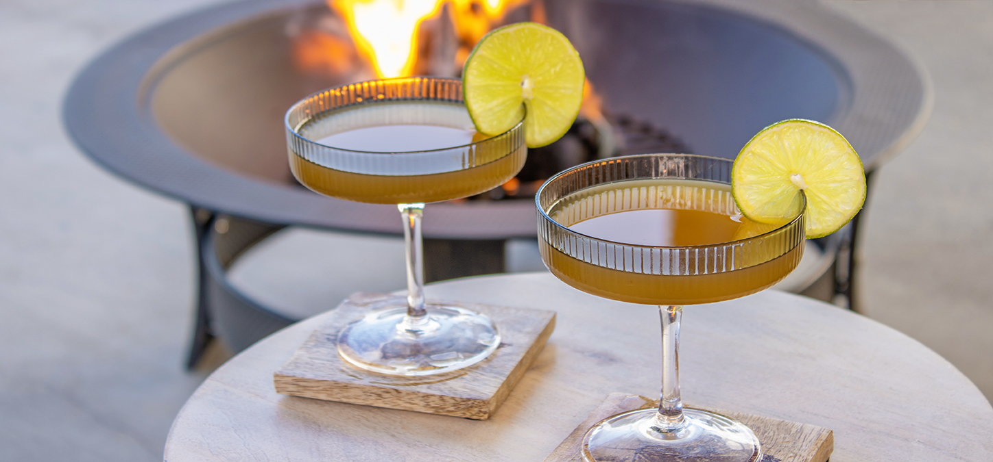 Two cocktails with lime garnish with a duraflame fire burning in an outdoor firepit in the background