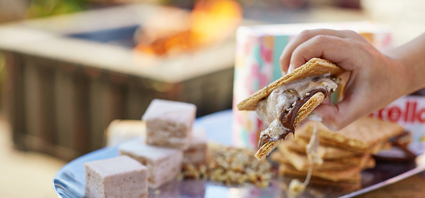 Hand holding a s'mores with ingredients and fire pit in background