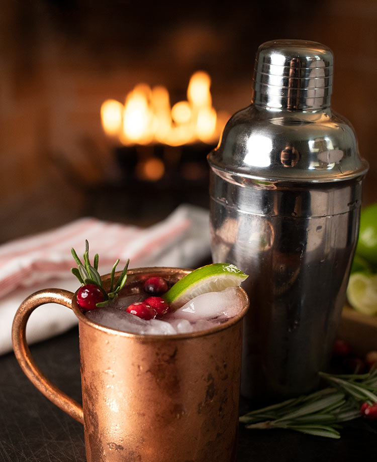 Libation in a tin mug with cranberry, rosemary and lime garnishes with blazing fire in a fireplace in the background