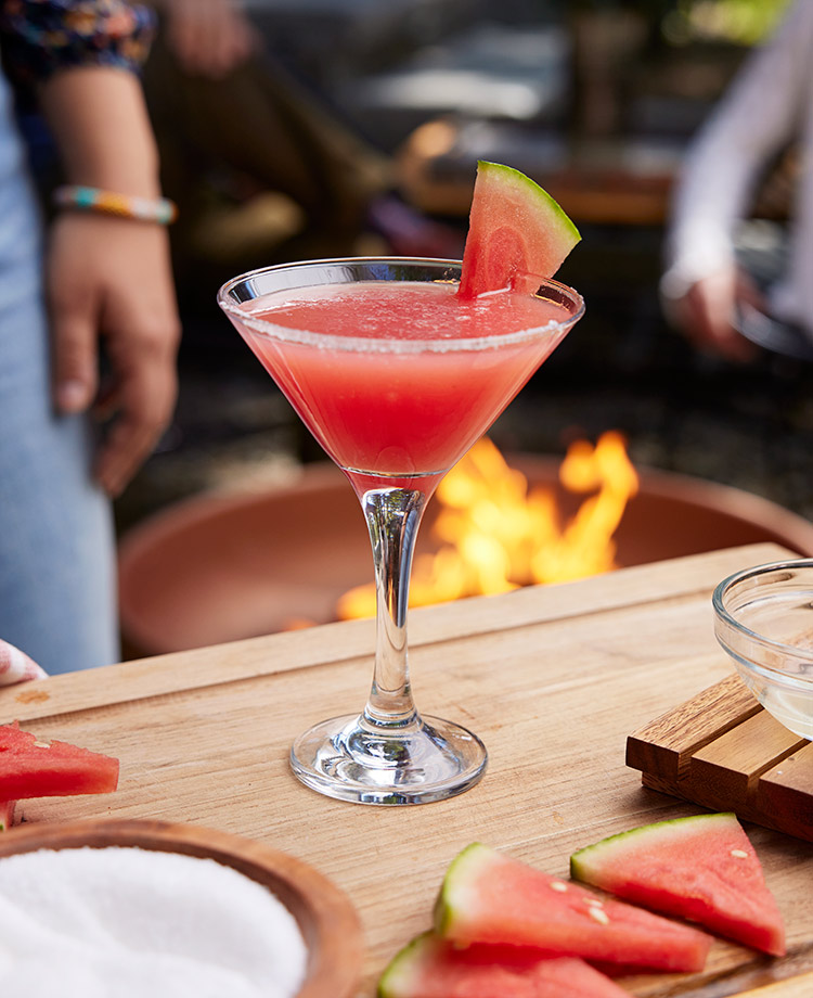 Libation in a martini glass with watermelon and lime, with blazing fire in an outdoor fire pit in the background