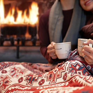 Couple cuddling under blanket holding mugs with duraflame firelog burning in fireplace in background