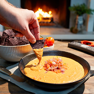 Hand dipping corn chip in spicy queso dip with a Duraflame fire burning in the background