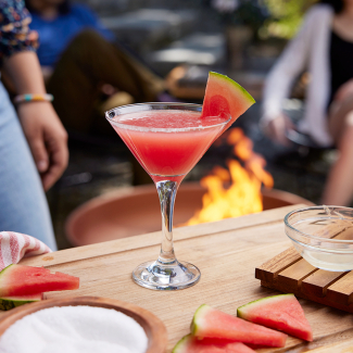 Libation in a martini glass with watermelon and lime, with blazing fire in an outdoor fire pit in the background